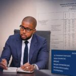 Illustration montrant un responsable assis à un bureau devant le drapeau haïtien, avec en arrière-plan un tableau des opérations financières de l’État et des éléments graphiques sur le financement du Trésor.