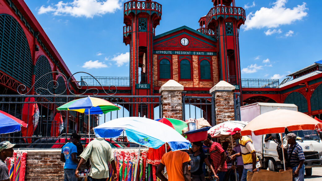 Scène animée devant un marché public emblématique en Haïti, avec des marchandes, des passants et des vendeurs de rue, illustrant le rôle central du petit commerce dans l’économie du pays.