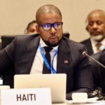 Le ministre haïtien de l’Économie et des Finances, Serge Gabriel Collin, prend la parole lors d’une réunion internationale, derrière une pancarte portant l’inscription « HAITI ».