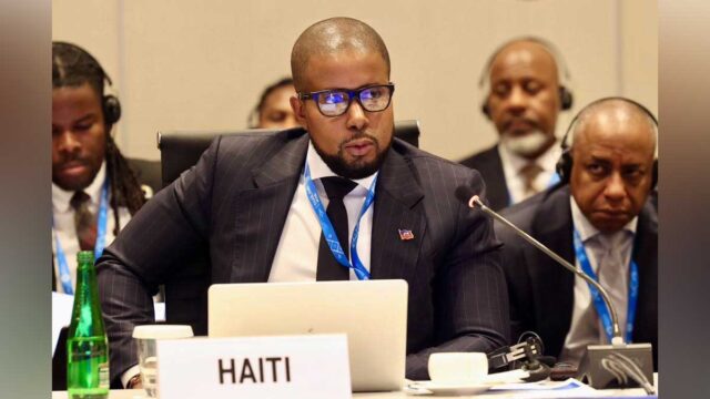 Le ministre haïtien de l’Économie et des Finances, Serge Gabriel Collin, prend la parole lors d’une réunion internationale, derrière une pancarte portant l’inscription « HAITI ».